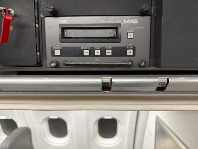 The image shows a Hi8 Video Cassette Player from MAS installed in what appears to be an airplane. The player has standard controls such as Power, Rewind/Play, Play/FF, Eject, and Stop. A handle labeled "PULL" is situated below the controls. The airplane interior, with its windows and seats, is visible in the background.
