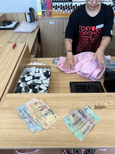 A retail transaction is observed at a store counter in Thành phố Kon Tum, Vietnam. An adult female, wearing a black t-shirt with red and white text (e.g., "TOKYOLIFE"), light-framed glasses, a wristwatch on her left wrist, and a bracelet and ring on her right hand, is positioned behind a light wood-grain counter. Her hands are placed over a light pink folded garment.
On the counter, various items are arranged:
* In the immediate foreground, two distinct stacks of Vietnamese Dong currency are present. On the left, a smaller stack consists of 20,000 VND (blue) and 10,000 VND (yellow/pink) notes. On the right, a larger, neatly folded or stacked bundle of 500,000 VND (teal) notes is visible, adjacent to a yellow rubber band and some dark liquid stains on the wood.
* To the left of the person's hands, a cardboard box is filled with numerous white and black plastic anti-theft security tags of various shapes. A crumpled white paper item is at the back of this box.
* Centrally located on the counter, between the person's hands and the box of tags, is a silver, dome-shaped magnetic security tag remover tool.
* To the person's right, a black rectangular device with a patterned surface, possibly a scanner or payment terminal, rests on the counter.
* Further to the left on the counter, a red utility knife or box cutter, a black tablet or clipboard with a cable, a clear plastic bag containing a white item, and a dark grey water bottle are present.
The background features light brown wooden cabinetry and shelving, with a display of small bottles, likely essential oils or fragrances, arranged on a tiered rack behind the person. A tall, decorative item with floral patterns is also visible. The scene depicts an ongoing or completed retail sale within a store environment.