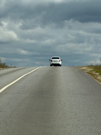 The image shows a white car parked on a road in what appears to be a rural area near Michurinsky, Russia. The sky is overcast with heavy clouds. The road is paved with a white line marking its edge. There is some vegetation along the side of the road. A road sign is faintly visible. The car is facing away from the camera.