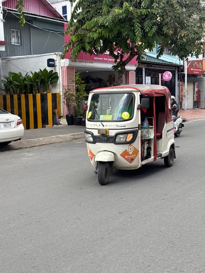In the city of Khan Boeng Keng Kang, Cambodia, a white and red auto-rickshaw, identified as a "TVS KING," is positioned on an asphalt road. The vehicle features an orange logo with the text "PassApp" and a yellow sticker with "PassApp" also visible on the windshield. A bottle of water is placed in a holder inside the auto-rickshaw. To the left of the auto-rickshaw, a white car with license plate "2BS-1776" is parked. Behind the parked car, a yellow and black striped barrier runs along a sidewalk. Further behind the barrier, lush green plants are situated in front of a building. The building has a gray exterior with a rolling metal door and a window with a metal grate. A pink-themed establishment with "DOBBASIR" signage is also visible. In the background, trees with dense green foliage are present, and other buildings and shops can be seen. To the right of the auto-rickshaw, a person wearing a helmet is riding a white motorcycle. The overall environment suggests an urban street scene with commercial establishments and a moderate level of pedestrian and vehicular activity.