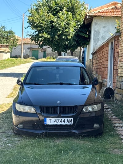 In Konop, Bulgaria, a black BMW sedan is parked on the side of a dirt road. The car is positioned in front of a narrow brick building and a lighter-colored structure with a red tile roof. A large tree with green foliage partially obscures the view of the buildings. The ground in front of the car is covered with green grass and some scattered stones, forming a low border along the road. The license plate of the car reads "T 4744 AM" with "BG" to the left and smaller text below. The sky is clear and blue.