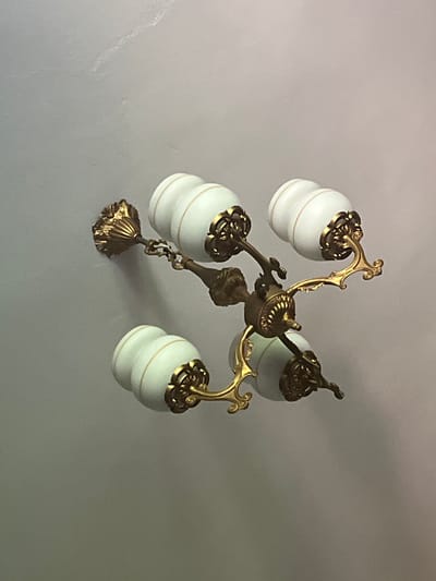 The image shows a close-up, angled view of a vintage brass chandelier with four frosted glass lampshades, each adorned with thin gold bands. The chandelier is attached to a plain, light grey ceiling. The lampshades are bulbous with a ribbed texture, tapering towards the top and bottom. Ornate brass fittings connect the lampshades to the central structure of the chandelier, featuring scrollwork and floral motifs. The central hub of the chandelier also displays decorative brass detailing. The lighting in the scene is soft, casting subtle shadows on the ceiling, suggesting it is either daytime with ambient light or the chandelier itself is not illuminated. The scene is devoid of any people or other objects. The location in the city of Ponebel, Ukraine, is not discernible from the image itself, as it solely depicts the interior of a room focused on the ceiling fixture.