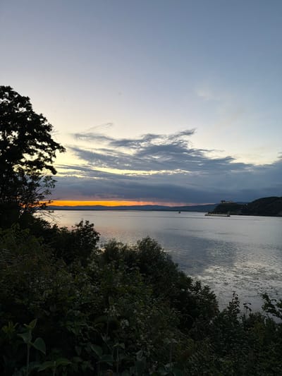 This is a photograph of a sunset over a body of water, likely the Danube River, with the Golubac Fortress visible in the distance on the right side of the frame. The foreground is dominated by dark green foliage and trees, obscuring much of the land. The sky transitions from a pale blue at the top to warmer orange and yellow hues near the horizon, with clouds scattered throughout. The water is calm with visible ripples reflecting the sky. A barge and a small boat are present on the water. The Golubac Fortress is perched on a hill overlooking the river, with some illuminated structures on its grounds. The scene appears to be a natural landscape with a historical landmark.