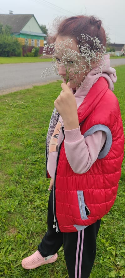 In an outdoor scene, a young person stands on a grassy area with a paved road and rural buildings in the background. The individual, viewed from the waist down, is wearing a red puffer vest over a pink hoodie and black sweatpants with pink stripes. Light pink Crocs are on their feet. They are holding a sprig of small white flowers in their right hand, bringing it towards their face, partially obscuring their features. The grassy area appears to be a yard or field, with some scattered debris. The buildings in the distance, including a house with a green roof and yellow and green accents, suggest a rural Belarusian setting, possibly within the territory of the Пацейкаўскі сельскі Савет. The sky is overcast.