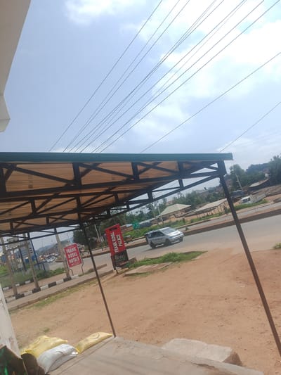 The image depicts an outdoor scene in what appears to be a roadside setting in Bukuru, Nigeria. A covered structure, likely a shelter or a stall with a corrugated roof supported by dark metal beams, dominates the foreground. Underneath the structure, there are bags of what looks like grain or fertilizer, suggesting a commercial or storage area.
To the left of the structure, there is a sign that reads "FRANK ROYAL HOTELS" and another sign with text that is difficult to decipher but appears to be in English. To the right, a red sign with white lettering states "FRANK ROYAL PALACE" and below it, "HOTEL", "RESTAURANT", and "BAR". These signs indicate the presence of hospitality businesses.
The scene includes a paved road with some traffic. A silver car is visible on the road. The background shows a mix of buildings, trees, and vegetation, indicative of a semi-urban or rural environment. The sky is partly cloudy, suggesting it is daytime. The overall atmosphere is that of a typical day in a Nigerian town, with elements of commerce and daily life. There are no people clearly visible engaged in any specific activity, but the presence of the road and businesses implies ongoing activity. The ground is a reddish-brown soil. The image is shot from a slightly elevated perspective, looking towards the road and beyond.