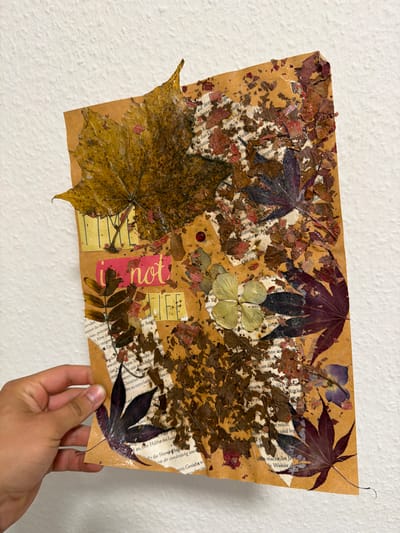 The image shows a collage of dried leaves and text, affixed to a piece of textured brown paper. The collage is held by a person's hand in the lower left corner. The collage is placed against a white, textured wall. The collage itself features a large yellow leaf at the top, with smaller dark red and purple leaves scattered throughout. Fragments of text from what appear to be book pages are visible beneath and around the leaves. Some text appears in yellow and pink strips, forming the words "TIME," "i not," and "LIFE." A small red bead is visible near the center of the collage. The collage is approximately 8.5 x 11 inches in size. The scene is indoors, with the white wall suggesting an interior setting. There is no specific indicator of the location within Nienburg/Weser, Germany.