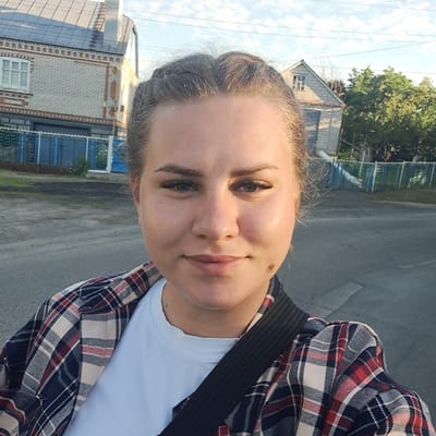 This media file shows a young woman taking a selfie outdoors. She is wearing a plaid shirt over a light blue t-shirt and has her hair pulled back. The woman has light skin, dark eyebrows, and is looking directly at the camera with a neutral expression. There is a small mole or mark on her cheek.

The background suggests a residential area. To the left, there are parts of buildings with brickwork and windows. A blue fence is visible. The ground appears to be an asphalt road. Trees are present in the background, and the sky is bright with some clouds, indicating it is daytime. The lighting suggests it could be morning or late afternoon.

The image has a slightly tilted perspective, common in selfies. There is no visible text in the image.