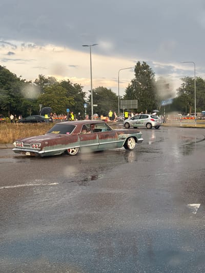 In Falkenberg, Sweden, a rusted and weathered light blue Chevrolet Impala is seen driving on a wet asphalt road. The car has custom red and white wheels. A person is visible in the driver's seat. Behind the Impala, a silver Peugeot 208 with the text "Nelle's" on its side is also on the road. There are several people gathered on the side of the road, some holding umbrellas, suggesting it may be raining or has recently rained. A black car is parked to the left. The environment appears to be a street with trees and streetlights. The sky is cloudy with hints of sunlight.