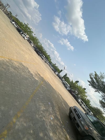 This image depicts a large, outdoor parking area or plaza in Ozuaha, Nigeria, viewed from a high-angle, diagonal perspective. The ground is paved with light-colored interlocking concrete blocks, primarily beige or light brown, with yellow painted lines marking parking spaces or boundaries visible on the darker grey section of the foreground. Numerous vehicles, including vans, SUVs, and cars of various models and colors (predominantly white, black, and grey), are parked in rows along the far left and central-right sides of the paved surface. A dark grey Nissan SUV is prominently parked in the lower right foreground. Green foliage from trees and shrubs lines the perimeter behind the parked vehicles, with taller, more distinct trees visible on the far right and left. Shadows from trees and parked vehicles are cast diagonally across the paved ground, indicating bright daylight conditions. The sky above is clear blue with scattered white cumulus clouds. A street light pole is visible among the parked cars on the right side. No individuals are present in the scene. Small pieces of white debris or litter are scattered on the paved surface in the foreground.
