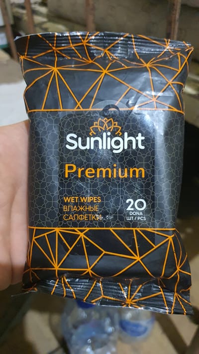The image shows a close-up of a black and orange package of Sunlight Premium wet wipes. The package has a geometric pattern of intersecting orange lines on a black background. The Sunlight logo, a stylized lotus flower in orange, is visible above the word "Sunlight" in white, sans-serif font. Below "Sunlight" is the word "Premium" in large, orange, serif font. Further down, "WET WIPES" is written in white, and below that, in a Cyrillic script, are the words "ВЛАЖНЫЕ" and "САЛФЕТКИ". To the right of the text, the number "20" is displayed in large white digits, with "DONA" and "ШТ/PCS" written below it in smaller text. The package is being held by a hand, partially visible on the left side of the frame. In the background, out of focus, are several plastic water bottles with blue caps. The image appears to be taken indoors, possibly in a shop or a home setting, with some blurred wooden structures visible in the upper background. There is no indication of the city of Karankul, Uzbekistan, in the image.