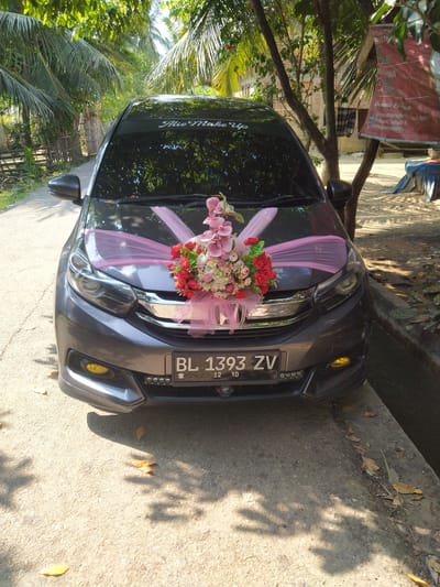 A dark gray car is parked on a paved surface, possibly a road or driveway, in an outdoor setting. The car is decorated with a pink ribbon bow and a floral arrangement on its hood. The license plate reads "BL 1393 ZV". The background features lush green foliage, including palm trees, and what appears to be a building constructed with red bricks. A red banner with text in Indonesian is visible hanging from a tree on the right side of the frame. The scene is illuminated by sunlight, casting shadows on the ground. The environment suggests a tropical or sub-tropical location, consistent with the city of Kruengbaroblangme, Indonesia.