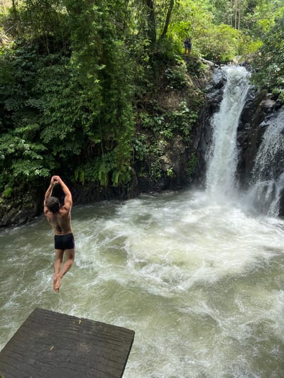 A shirtless Caucasian male, seen from the back, wearing black swim briefs, is suspended mid-air over a turbulent, frothy water body. He holds a rope swing with both hands raised above his head. Below him, to the lower-left, a dark, striped wooden platform is partially visible. To the right, a powerful waterfall cascades white water down dark rock formations into the agitated pool. Dense, vibrant green tropical vegetation covers the steep banks surrounding the water. In the upper background, two additional individuals are present: one person is standing on a rocky ledge above the waterfall, and another is partially visible near the top of the waterfall. This scene is identified as Aling-Aling Waterfall in Ambengan, Indonesia.