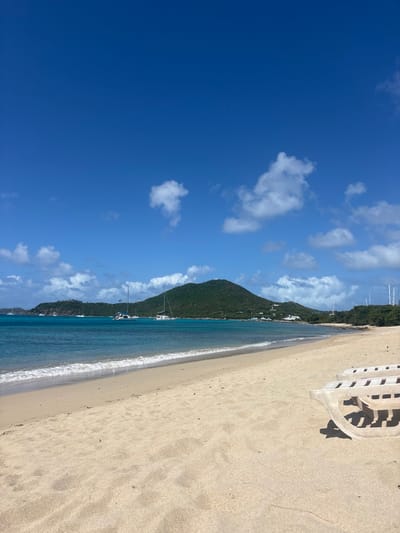 The scene depicts a sunny beach located in Spanish Town, British Virgin Islands. The foreground features light tan, fine-grained sand occupying the lower half of the image. The arm and partial backrest of a light-colored, possibly beige, plastic beach lounge chair are visible on the far right. A narrow strip of white, foamy water marks the breaking waves at the sandy shore. The midground presents a body of calm, clear turquoise-blue ocean water. Several sailboats and catamarans are anchored in the bay, with two larger vessels visible in the central-left. The background is dominated by lush, green, tree-covered hills rising from the ocean, extending across the horizon. Scattered white structures are discernible along the lower slopes of these hills. The sky is bright blue with scattered white cumulus clouds. No people or animals are present.