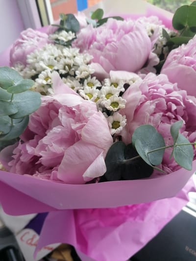 A close-up, vertical, medium shot captures a bouquet of pink peonies and white daisies. The bouquet is partially wrapped in bright pink paper, with some of the paper folded and creased. Several green eucalyptus leaves are interspersed throughout the flowers. The background is blurred, with hints of a window frame and what appears to be a brick wall, suggesting an indoor setting. There are no people visible in the image. The lighting is soft, coming from the right side of the frame. The bouquet appears to be resting on a surface, though it is mostly obscured by the wrapping paper.