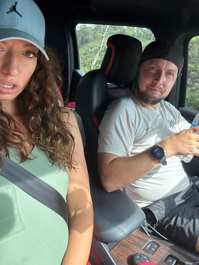 A man and a woman are inside a vehicle, possibly a car or SUV, in an unidentified location, presumed to be in the Suối Giàng, Vietnam. The man, with a beard, wearing a black beanie, and a light-colored short-sleeved shirt with a logo, is reclining in the passenger seat. He's holding a plastic water bottle in his left hand and wearing a smartwatch on his left wrist. He's positioned in the upper half of the image. The woman, wearing a light blue baseball cap with a logo, has long curly brown hair. She is wearing a light green shirt and a seatbelt, and she is positioned in the lower half of the image. The vehicle's interior features dark leather seats with red accents and wood-grain trim on the console. The vehicle's sunroof reveals trees and foliage overhead.