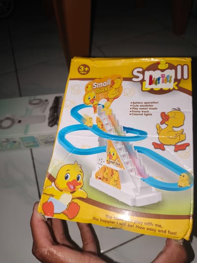 A toy product box is held by a person's hand. The box is yellow and features cartoon illustrations of ducks and a slide. The text "Small Duck" is prominently displayed in various locations on the box. There are also bullet points listing features such as "Battery operation," "Cute escalator," "Play sweet music," "Funny track," and "Colored lights." The box appears to be made of cardboard and is sealed with plastic. The background shows a blurred indoor environment with tiled flooring and furniture. The image is taken from a close-up perspective, focusing on the toy box and the hand holding it.