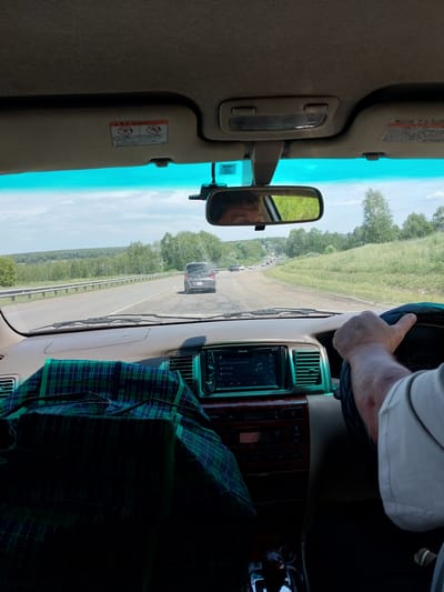 The image is taken from inside a car traveling on a road in Nalobikha, Russia. The perspective is from the driver's seat, looking forward and slightly to the right. The road is asphalt and appears to be in good condition, with a guardrail on the left side. There are other cars on the road in the distance. The landscape on either side of the road is rural, with green fields and trees under a cloudy sky.
Inside the car, the dashboard is visible, with a navigation system and various controls. A green and blue plaid blanket is draped over the passenger seat. The driver's hand is on the steering wheel. A side mirror reflects the driver's face and the passing scenery. The sun visors are up, and one has a warning sticker with Japanese text and symbols. The overall impression is of a road trip on a summer day.
