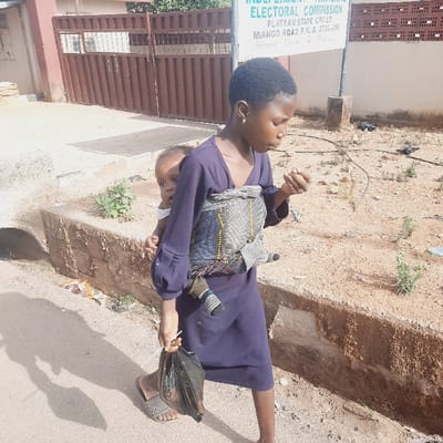 The image shows a young woman in a blue dress carrying a toddler on her back in a sling. She is walking along a dirt road with a plastic bag in her hand. The woman is looking down at the ground and the toddler is looking at the camera. The scene takes place in Kufang, Nigeria, which is located at latitude 9.863 and longitude 8.867. There is a signpost in the background displaying "Independent Electoral Commission Plateau State Office". There are some plants growing alongside the road and a concrete barrier separating the road from the sidewalk. The road is relatively empty. The woman is wearing a blue dress with a white and black pattern. The toddler is wearing a white t-shirt and blue pants. The plastic bag she is carrying is black and appears to be filled with items.