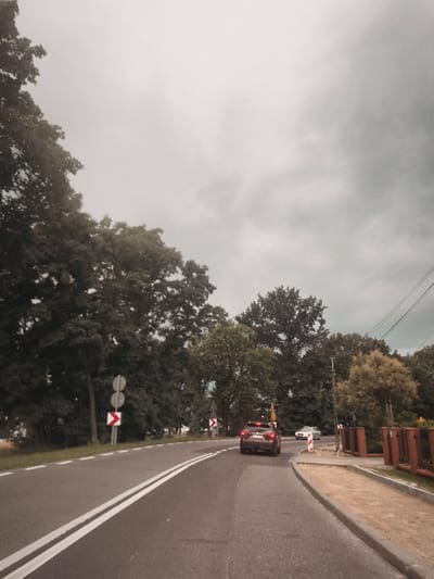 The image depicts a two-lane asphalt road curving to the right, bordered by trees and a sidewalk. The road has double white lines in the center and white dashed lines on the right edge. Two cars are visible: a dark-colored SUV in the foreground, traveling away from the viewer, and a silver sedan further ahead, also moving away. Traffic signs, including a chevron sign indicating a curve, are present along the roadside. The sky is overcast with heavy clouds. The scene appears to be in Olszewnica Nowa, Poland, based on the presence of a Polish license plate on the SUV. A brick fence and an area of disturbed sand are visible on the right side of the road. Electrical wires are strung overhead.