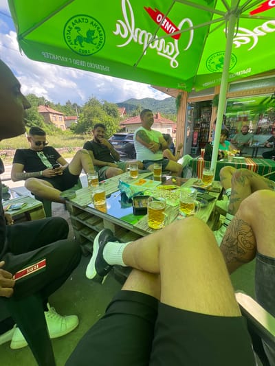 This image depicts an outdoor social gathering at a casual establishment in Касапите, Bulgaria, on a clear, partly cloudy day. The scene is framed by two large, green promotional umbrellas. One umbrella prominently displays the Cyrillic text "СТАРА ЗАГОРА" (Stara Zagora), "БЪЛГАРСКО ПИВО С ХАРАКТЕР" (Bulgarian Beer with Character), and a lion/cat logo with the year "1892". The other umbrella features "ЛИНФО" (LINFO) branding.

Approximately eight to ten adults, primarily males, are present. In the immediate foreground, the lower half of two individuals is visible: one male wears a black tracksuit with a red and green "ICON" stripe and white sneakers; another male, with tattooed legs, wears black shorts, white socks, and black Nike sneakers.

Around a rustic table constructed from wooden pallets, several men are seated. One man in a black t-shirt and sunglasses is focused on his smartphone. Another bearded man in a dark green t-shirt is smiling. A third man, with arm tattoos, wears a white t-shirt. Most individuals are dressed in shorts and short-sleeved shirts, indicating warm weather. The table holds multiple glass mugs filled with a light-colored liquid (consistent with beer), bowls containing snacks such as chips and pretzels, and personal items like smartphones and an ashtray.

In the mid-ground, a storefront, likely a small bar or café, with glass doors and windows, reveals shelves stocked with bottles inside. An older woman in a green top and two additional men are visible within the establishment. Beyond, the background features rural houses with red-tiled roofs, dense green foliage, and distant hills or mountains under a partly cloudy blue sky. A dark-colored SUV is partially visible parked behind the group. The overall ambiance is relaxed and social.