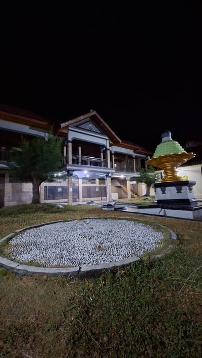 This is a medium-shot, eye-level, vertical, nighttime photograph of a building and its exterior surroundings in Lhokdagang, Indonesia. The building appears to be two stories with a dark roof and light-colored walls. It has a covered balcony area on the second floor with a railing. Decorative architectural elements are visible on the facade. In the foreground, there is a circular garden bed filled with small, light-colored stones, bordered by a concrete edge. To the right of the stone garden, there is a dark, ornate fountain or monument with a green, layered top. The ground is covered in sparse grass and some dry vegetation. The lighting in the scene appears to be artificial, possibly from streetlights or the building itself, creating shadows and highlighting certain features. No people are visible in the image. The overall impression is one of a quiet, well-maintained public space at night.