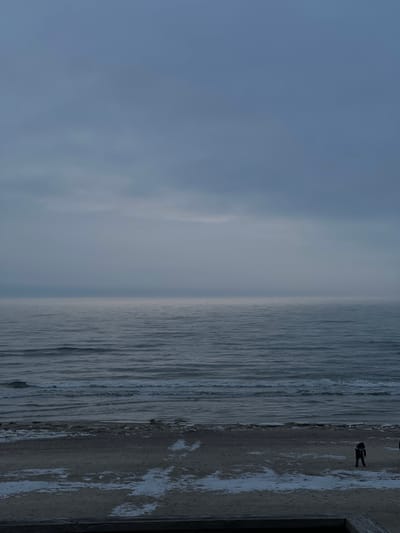 The image depicts a vast coastal scene in Morskoe, Russia, under an overcast sky. The upper two-thirds of the frame are dominated by a pale grey-blue sky, merging indistinctly with a hazy horizon line. Below the horizon, a wide expanse of choppy, dark grey-blue water shows distinct wave patterns approaching the shore. The bottom third of the image reveals a sandy or gravel beach, partially covered with irregular patches of white snow or ice. Along the immediate water's edge, broken ice or debris is visible. Two human figures are present on the beach, both dressed in dark, heavy clothing. One individual stands center-right, facing generally towards the sea. A second individual is partially visible on the far right edge of the frame. In the extreme foreground, a dark wooden structure, possibly a railing or ledge, spans the bottom of the image, indicating an elevated perspective. The overall environment suggests a cold, possibly winter, day.