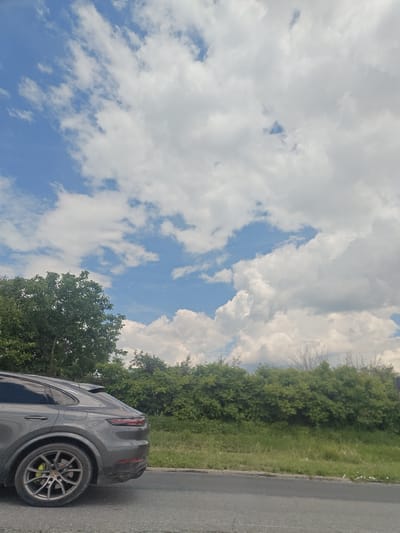 The image captures a daytime scene in Vadul lui Vodă, Moldova, featuring a dark gray SUV parked on the right side of an asphalt road. The SUV, viewed from its rear quarter panel, shows a spoiler and tinted windows. The immediate foreground consists of the road, followed by a strip of grass and a dense line of green trees and bushes that form a natural barrier. Above the foliage, a vast expanse of sky dominates the upper portion of the frame, characterized by a mosaic of blue sky and fluffy white cumulus clouds. The lighting suggests it is a bright day, with the sun likely overhead or slightly to the side, illuminating the clouds and the landscape. The setting appears to be an area bordering a green space or park within the city.