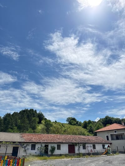 The image presents a view of Ogradna, Bulgaria, with the sky occupying a dominant portion. The sky is blue with streaks of white clouds. A building with a red tiled roof is visible on the right side of the image, nestled on a hillside covered in greenery. Some sort of play structure can be seen on the right.