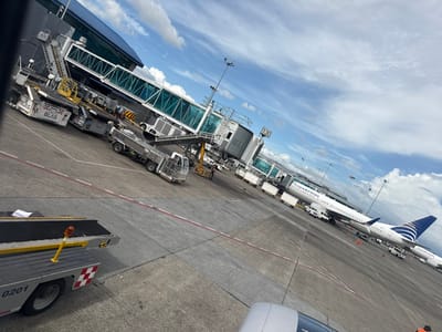 An airport tarmac is visible from an elevated angle. A Copa Airlines plane sits at a gate, connected by a jet bridge. Various airport vehicles and equipment surround the plane and gate. A portion of an airplane wing is visible in the upper left corner of the image. The sky is partly cloudy.