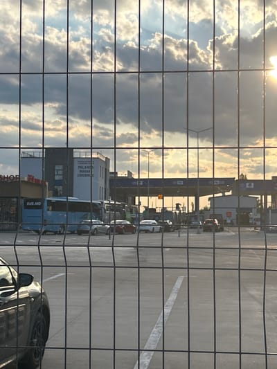 The image shows a view of the Palanca, Moldova border crossing through a wire fence. The sky is filled with dramatic clouds, with the sun shining brightly on the right side of the frame, casting a warm glow. In the middle ground, several vehicles are visible, including a blue bus with "SVS.MD" written on its side, and cars lined up near what appears to be a border control building or customs station. The architecture suggests a functional, modern facility. The foreground is dominated by the metal grid of the fence.
