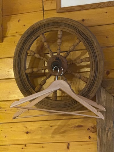 An antique wooden steering wheel, with a metal hook, hangs on a textured yellow wooden wall alongside two wooden coat hangers. The location is identified as the city of Ketoshi, Russia.