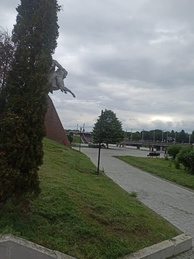 The image depicts an outdoor scene in Nazran, Russia, featuring a large equestrian statue partially obscured by a tall evergreen tree in the foreground. The statue, made of a light-colored material, shows a rider on a horse, positioned on a triangular, reddish-brown monument base. A grassy, sloped area surrounds the base. To the right of the grassy slope, a paved walkway with a gray, brick-like pattern runs diagonally through the frame. Beyond the walkway, there is a green lawn with benches and more trees. In the background, a body of water, possibly a river, is visible, crossed by a bridge. Several people are present in the midground, walking on the pathway or sitting on benches. There are also buses visible on the other side of the water, near a set of stairs leading upwards. The sky is overcast with heavy, gray clouds. The overall impression is a public park or memorial area on a cloudy day.