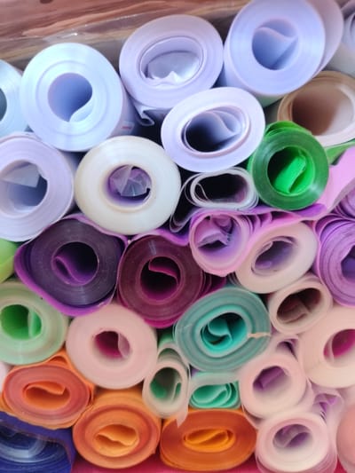 This image displays a tightly packed collection of fabric rolls, likely of various colors and textures. The rolls are stacked vertically and horizontally, creating a textured and colorful composition. The colors range from light purples and whites at the top, transitioning to darker purples, greens, blues, and oranges towards the bottom. Some rolls appear to be made of sheer or translucent material, while others have a more opaque finish. The lighting is soft and diffused, suggesting an indoor environment, possibly a retail space or storage area. There are no people or animals visible in the image, and no specific actions are being performed. The image does not provide any information or context about the city of Kalach, Russia, or its specific locations.