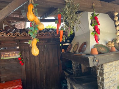 The image depicts an outdoor covered area, likely a patio or porch, with rustic wooden beams and a tiled roof. The scene is decorated with hanging dried herbs, strings of garlic, and several artificial vegetables including bright orange pumpkins, red peppers, and carrots. On the left side, a wooden door is visible, adorned with a sign in Bulgarian that reads "Лятна градина" (Summer Garden). Below the sign, a small bunch of red peppers hangs. To the right, a brick oven or fireplace structure is present, with decorative plates and terracotta pots placed on the mantle. The overall ambiance suggests a traditional, perhaps seasonal, outdoor setting in the village of Zheravna, Bulgaria, known for its preserved architecture and folk traditions. No people are present in the image.