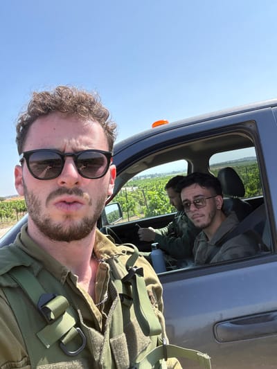 The image shows three men standing outside a gray pickup truck in a vineyard. The man in the foreground is wearing a green military uniform, black sunglasses, and a beard. He is looking at the camera. Behind him are two other men sitting in the truck, looking at the camera. The man on the driver’s side is wearing green clothing and a beard. The man on the passenger side is wearing a green military uniform and has a beard and short dark hair. The truck has a roll-up window on the passenger side, with a small yellow light on the top. The man in the foreground is holding a military backpack. The image was taken near the vineyards outside Mazkeret Batya, Israel, at 31.824 N, 34.842 E. The background is a vineyard with a clear blue sky. The scene appears to be a casual encounter between three friends.