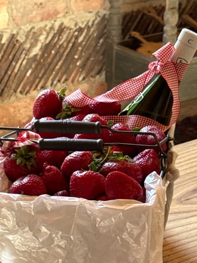 In Alagir, Russia, a dark metal basket filled with ripe strawberries and lined with white crumpled paper sits on a wooden surface. The basket has two dark handles. A green bottle, possibly containing champagne or wine, is placed among the strawberries and is adorned with a red and white checkered ribbon tied around its neck. The background features a rustic brick wall with wooden planks.