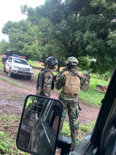 The scene depicts a rural or semi-rural setting in Mkavior, Nigeria, characterized by a muddy dirt road bordered by dense green foliage under an overcast sky. Two individuals in military-style camouflage uniforms, tactical vests, and helmets stand centrally on the road. The individual on the left wears a black tactical vest and helmet, holding a rifle, and faces the individual on the right. The individual on the right wears a tan tactical vest and helmet, also appearing to hold a rifle, and faces away from the camera. Both individuals are engaged in an interaction, possibly a discussion or briefing.

In the background, to the left, a white Toyota Hilux-style pickup truck is parked on the muddy road, equipped with a roof rack and blue and red emergency lights on its roof. Further back on the right side of the road, several motorcycles are parked in the grassy area. A third individual, also in camouflage, is partially visible in the side mirror of the vehicle from which the image was captured, which is dark-colored. The ground is uneven, indicating recent moisture. Distant structures or walls are faintly visible amidst the trees to the left.
