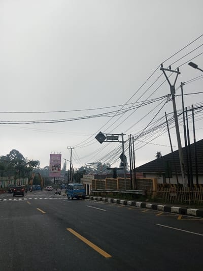 The image captures a detailed street scene under an overcast sky, likely in an urban or semi-urban area of Indonesia, suggested by the visible text and vehicle type. The time of day appears to be daytime, but with diffused light due to the heavy cloud cover.
In the foreground, a dark asphalt road dominates the lower portion of the image. It features clear yellow double solid lines, indicating a no-passing zone, and a single dashed yellow line, likely for a turn lane or parking. On the right side of the road, a black and white striped curb with yellow markings suggests a restricted parking or stopping area. A small, light blue van, possibly an angkot (public minivan), is stopped or parked adjacent to this curb.
Further down the road, in the midground, several vehicles are visible. A red car is prominent on the left side of the road, with other cars and motorbikes visible behind it, suggesting active traffic flow or an intersection. To the far left, a partially visible green and white sign reads "PUNCAK" followed by obscured text like "MENUJU..." and "TERBUKA UNTUK". This strongly suggests the location is near the Puncak area in West Java, Indonesia. Behind this sign and the vehicles, dense green foliage indicates a treed or hilly landscape.
A large, brightly lit billboard stands tall on the left side of the road, featuring an advertisement for "GoodDay" coffee, with the words "Kopi Gaul" also visible below it. The billboard depicts several individuals, likely enjoying the beverage. Below the billboard and partially obscured by trees, some construction equipment, possibly an excavator or crane, can be seen.
The midground and right side of the image are characterized by a dense network of overhead utility wires and numerous utility poles. One prominent pole near the center of the image supports two diamond-shaped road signs, one directly above the other, though their specific meaning is not clear from the angle. Another large utility pole with multiple cross arms and wires stands on the far right.
Behind the utility poles and wires on the right, there is a building with a dark, possibly tiled, roof. A fence made of light-colored blocks and wood runs along the property line, separating the building from the road. Adjacent to the blue van, a small, light-colored building or guard post is visible. The background shows more distant hills or elevated terrain covered in trees, blending into the consistently overcast sky.
No specific activity beyond general traffic movement is evident, and no distinct emotions are discernible from the scene. The image presents a typical, bustling road environment in a Southeast Asian setting.