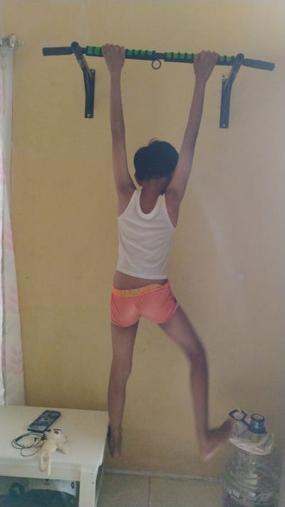 A person is hanging from a pull-up bar mounted on a yellow wall in Banjarsari, Indonesia. The person is wearing a white tank top and orange shorts, with their arms extended and hands gripping the bar. Their legs are dangling, and their body is blurred due to motion. Below the pull-up bar, a white table is visible on the left, with a mobile phone, headphones, and a stuffed toy on it. To the right, a large plastic water jug is present. The environment appears to be indoors, with a curtain on the far left and tiled flooring.