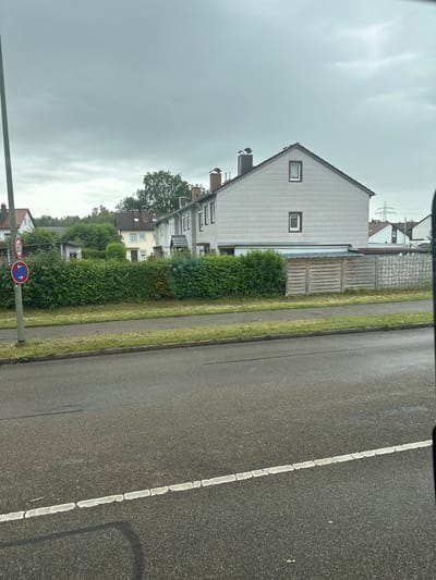 The image shows a residential street in Memmingen, Germany. The scene is captured from a slightly elevated perspective, looking across a road towards a row of houses and a dense green hedge. The road in the foreground is asphalt and appears wet, with a white dashed line marking the lane. To the left of the road, a tall wooden pole supports two circular traffic signs: one red with a white horizontal bar indicating "no stopping," and another red circle with a white arrow indicating a "turn" or "direction" restriction. Behind the hedge, several houses are visible, characterized by their pitched roofs and light-colored facades. One house in the center is larger and appears to be a two-story dwelling with a gray facade and red-trimmed windows. Another house to the left is yellow with dark-trimmed windows and a red roof. The sky is overcast with heavy gray clouds. Behind the houses, trees and vegetation create a natural backdrop. A wooden fence separates the properties from a grassy area. The overall impression is one of a typical suburban street on a cloudy day.