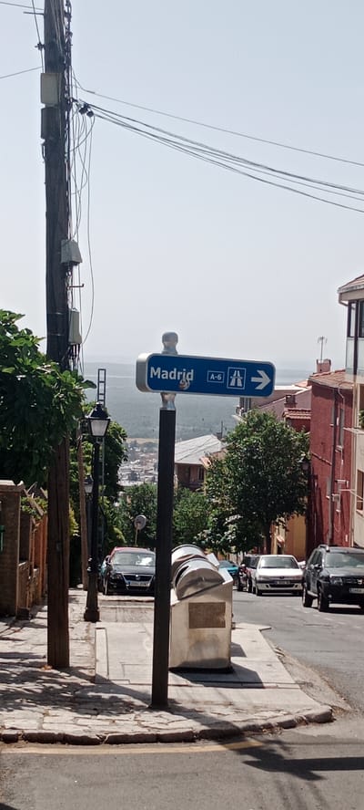 The image captures a street scene in San Lorenzo de El Escorial, Spain, dominated by a directional sign indicating "Madrid" with the A-6 highway symbol and an arrow pointing to the right. The sign is mounted on a dark pole. To the left, a tall wooden utility pole is laden with numerous electrical wires and boxes, partially obscured by green foliage. A traditional-style street lamp is also visible on the left side of the street.

The street itself is paved with asphalt, with a sidewalk made of concrete and stone slabs. Several cars are parked or driving on the street, including a black car with a visible license plate, a white car, and a dark SUV. Buildings with red and beige facades line the right side of the street, suggesting a residential or commercial area. In the background, a hazy vista of hills and trees can be seen, indicating the town's setting within a more rural landscape. The sky is clear and bright, suggesting a sunny day.
