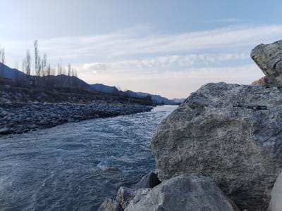 The scene depicts a riverine landscape in Qoraqiya, Uzbekistan. The foreground on the right is dominated by several large, grey, textured boulders. A flowing river with bluish-grey, moderately choppy water occupies the left and central midground, extending towards the distant horizon. The river's left bank is densely covered with numerous rounded grey river stones. Beyond this bank, an elevated slope features sparse vegetation, including several tall, slender, bare deciduous trees. A dark, indistinct structure is partially visible behind the trees on the far left. In the background, a range of dark, undulating mountains follows the river's course under a light blue sky with scattered wispy white clouds. No persons, animals, or artificial conveyances are present.