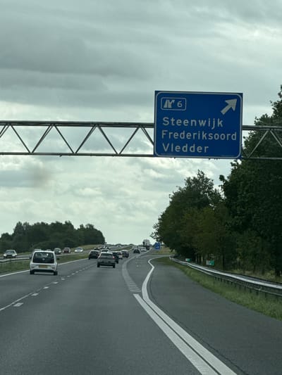 The image depicts a highway scene on a cloudy day. A blue overhead road sign displays directions for "Steenwijk," "Frederiksoord," and "Vledder," with an arrow indicating the direction for these locations. The sign also shows "A6" and a smaller symbol. Numerous cars are driving on the multi-lane highway. To the right of the highway, there is a guardrail and dense green trees. On the left side of the highway, there are more trees and a slightly elevated embankment. The road appears to be in or near the city of Tuk, Netherlands, given the destination names on the sign. The weather is overcast, with a gray sky.