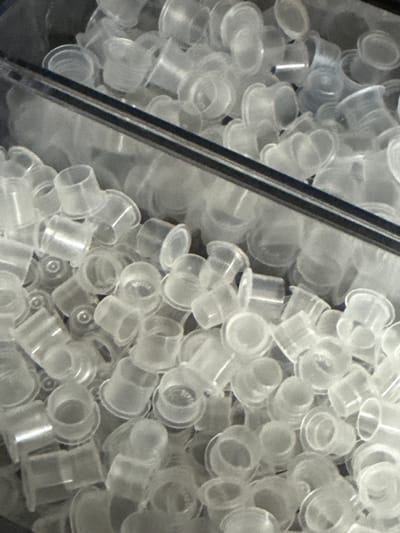 This is a close-up, overhead shot of a large quantity of small, clear plastic cups. The cups are stacked and piled together, filling the frame. They appear to be disposable, possibly ink caps used for tattooing or pigments for art. The lighting is somewhat diffuse, creating soft shadows and reflections on the plastic surfaces. The background is dark and out of focus. There is a dark, vertical object with a subtle metallic sheen running along the left edge of the image, which could be part of a container or shelving. The image is likely taken indoors, given the controlled lighting and the packed nature of the objects. There is no visible text in the image.