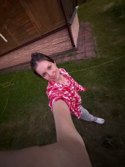 A young woman is taking a selfie outdoors in an overcast, twilight setting. She is wearing a red and white checkered shirt, gray sweatpants, and white crocs. She is standing on a grassy area next to a wooden structure with a sloped, thatched roof. The ground is covered in grass and some scattered bricks. The woman is looking at the camera with a slight smile. The scene suggests a casual, domestic setting, possibly a backyard in a rural or suburban area. The lighting indicates it is either dawn or dusk. There is no visible text in the image. The location context provided is Кривандино, Russia.