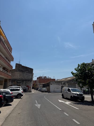 This is a medium-angle, outdoor, vertical shot of a street in Vilanova I La Geltrú, Spain, taken on a sunny day. The image is dominated by a clear blue sky, with a few wispy clouds. The street is lined with buildings, some appearing older and weathered, others more modern.
Several cars are parked along the street. In the foreground, a white van with "Correos Express" visible on its side is parked. Further down the street, other cars, including a white SUV, are visible. A tree with green foliage partially obscures the view of some buildings. There is graffiti on a wall in the middle ground. The street itself has white lane markings, including a large arrow indicating a direction of travel.
The time of day appears to be late morning or early afternoon, given the bright sunlight and the shadows cast. The weather is fair and warm. The scene depicts a typical urban streetscape with parked vehicles and residential or commercial buildings.