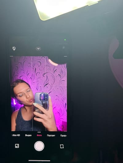 The image shows a young woman taking a selfie with her smartphone. She is holding the phone in her right hand, and the phone's screen displays a grid overlay, indicating it is in camera mode. The woman has her lips pursed in a kiss-like gesture, and her eyes are looking at the phone's camera. She is wearing a dark tank top, and her nails are painted a light green color. The background is a wall with purple floral wallpaper and what appears to be a ring light providing illumination. The setting is domestic and appears to be indoors. There is no information in the image to suggest it is located in Bishkek, Kyrgyzstan.