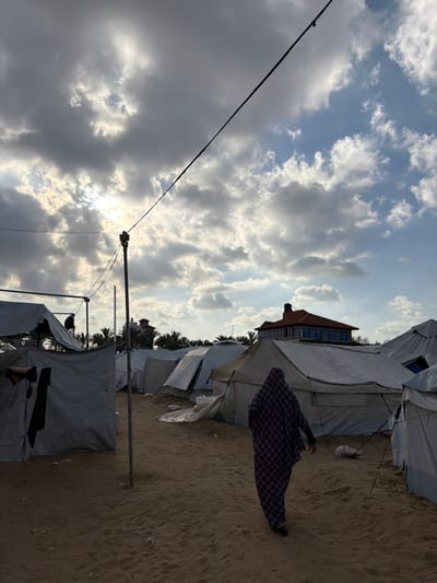 The image depicts an outdoor scene in a-Zawayda, Palestinian Territory, characterized by a sandy ground cover under a sky with extensive cloud cover and areas of visible sunlight. A temporary encampment is present, featuring numerous white and light-colored canvas tents of varying sizes, arranged across the mid-ground and foreground. A single individual, observed from behind, walks away from the camera towards the right side of the frame, clad in a full-length, patterned purple and black garment with a head covering. In the far background, beyond a line of palm trees and tent structures, a multi-story building with a reddish-brown tiled roof and reflective blue windows is discernible. An overhead utility cable, dark in color, traverses the upper portion of the frame diagonally, connecting to a vertical metal support pole situated in the left-center. Dark clothing items are visible hanging on a line or structure near a tent in the left foreground. Minimal scattered debris is present on the sandy surface.