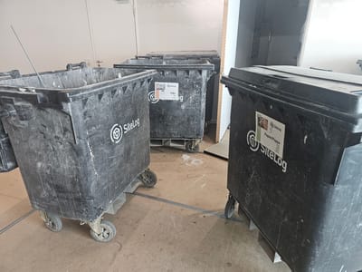The image shows several large, black, wheeled bins, likely for waste disposal. These bins are positioned on a concrete floor in what appears to be an indoor space, possibly a construction site or storage area, given the general disarray and unfinished elements in the background. One bin in the foreground on the right has a label that reads "SiteLog Papier/Pappe" and a smaller label with a barcode and text. Another bin in the middle left also has a "SiteLog" logo. The bins are dirty and show signs of wear and tear. The overall lighting is neutral, suggesting an indoor setting with artificial light or diffused daylight. No people are visible in the image. There is no specific information in the image to indicate a location in Darmstadt, Germany.