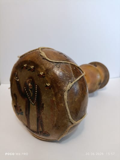 The image displays a handcrafted drum-like object with painted figures on its surface. The object is made of a porous material, possibly animal skin stretched over a gourd, and is bound with twine. The painting depicts two stylized human figures with intricate patterns and what appear to be decorative elements or clothing. Several small, bird-like figures are also painted around the main figures. The object rests on a white surface, likely a table or floor, with a plain white background. The lighting suggests an indoor setting. The timestamp at the bottom right of the image indicates "20.06.2026 15:57", and the bottom left corner displays "POCO M6 PRO". There is no indication of the city of Bender, Moldova, or any specific urban environment within the image itself.